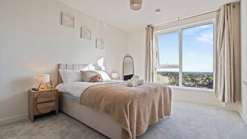 A bedroom with a large window, a bed with a fluffy blanket, a bedside table with a lamp, and a view of a cityscape.