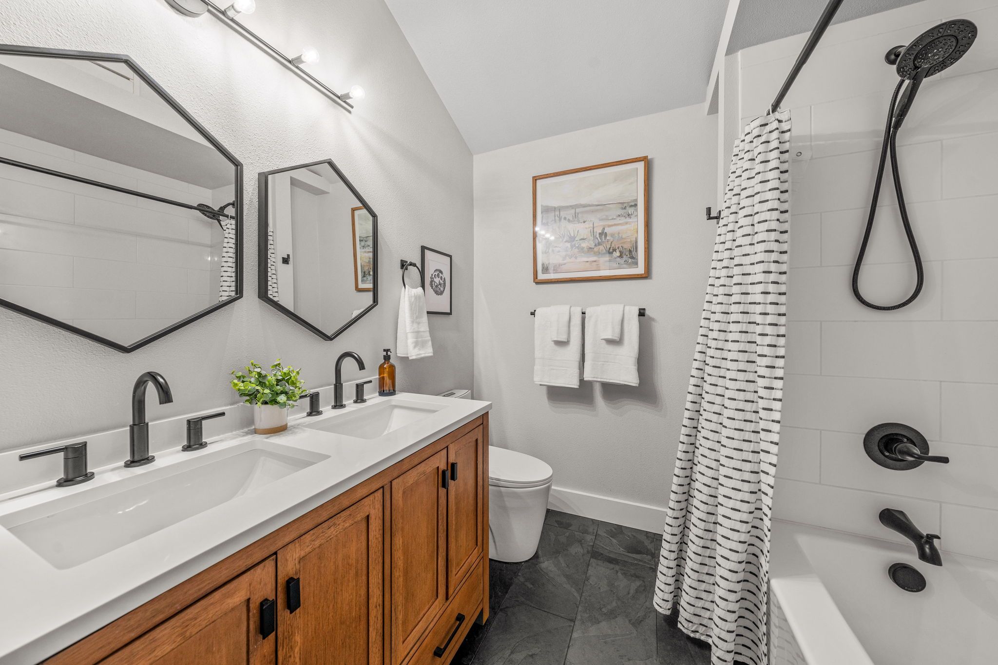 A modern bathroom with a large mirror, dual sinks, wooden cabinets, a toilet, a shower with a black hose, and a framed picture on the wall.