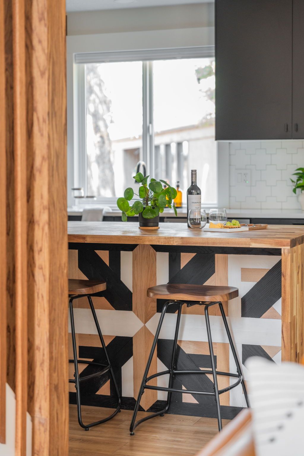 A kitchen with a wooden island, two stools, a potted plant, and a bottle of wine.