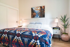 Un dormitorio con cama, mesita de noche, lámpara, planta en maceta y un cuadro en la pared.