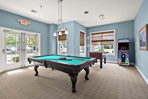 Une table de billard dans une pièce avec un mur bleu, une porte avec un panneau indiquant la sortie et une borne d’arcade.