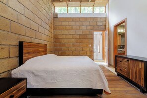 A bedroom with a brick wall, a wooden headboard, a large bed with a white quilt, a wooden dresser, and a view of a bathroom through an open door. - Heated Indoor Pool, Fire Pit, Billards. (Valle de Bravo)