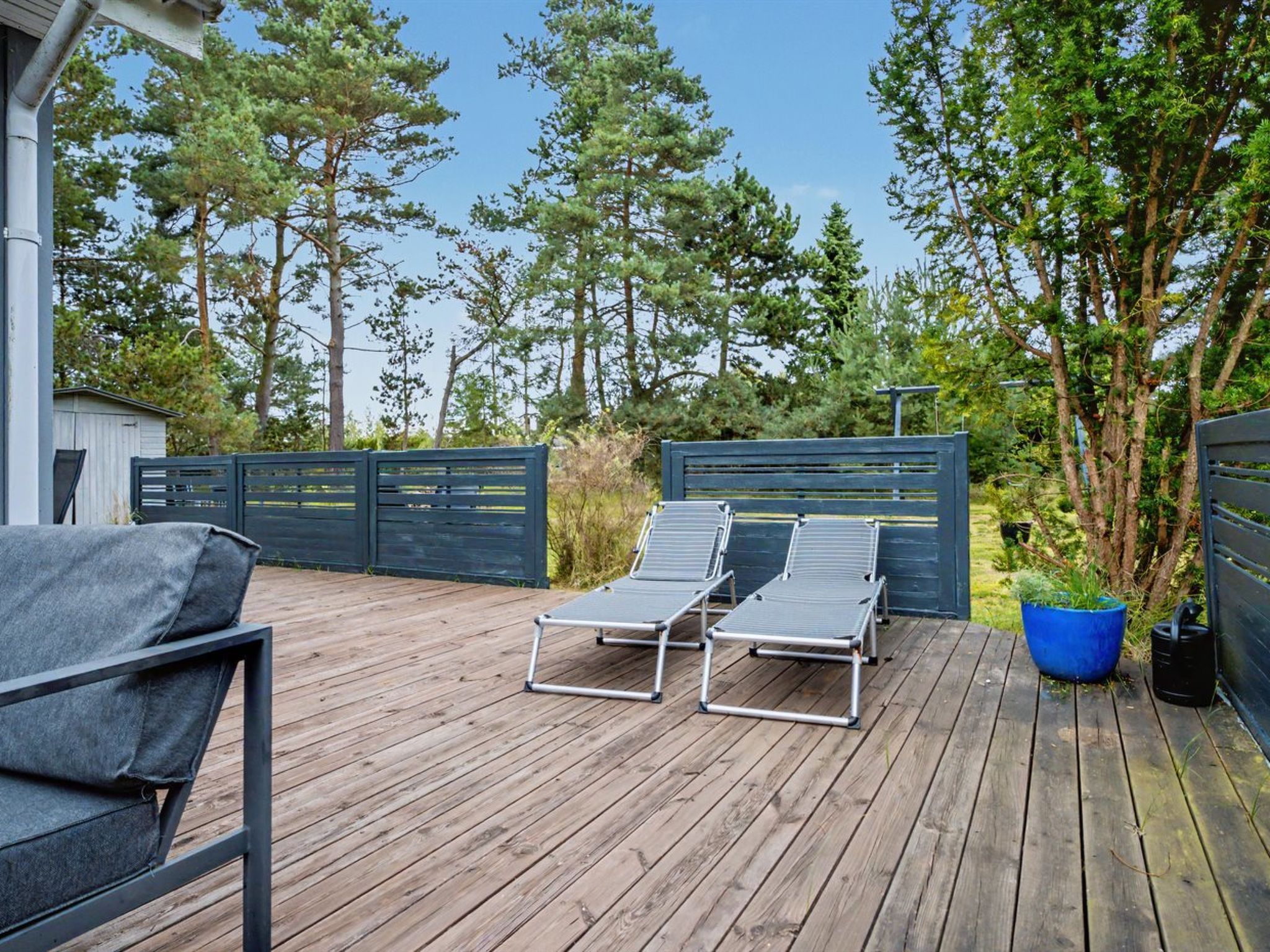A wooden deck with two lounge chairs and a small table, surrounded by a metal fence and trees.