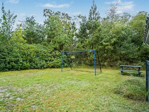 Ein Spielplatz mit Schaukeln und einem Picknicktisch in einer grünen, von Bäumen umgebenen Fläche.