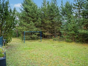 Ein grünes Außengelände mit einer blauen Schaukel und einer Spielkonstruktion mit Netz, umgeben von dichten Bäumen.