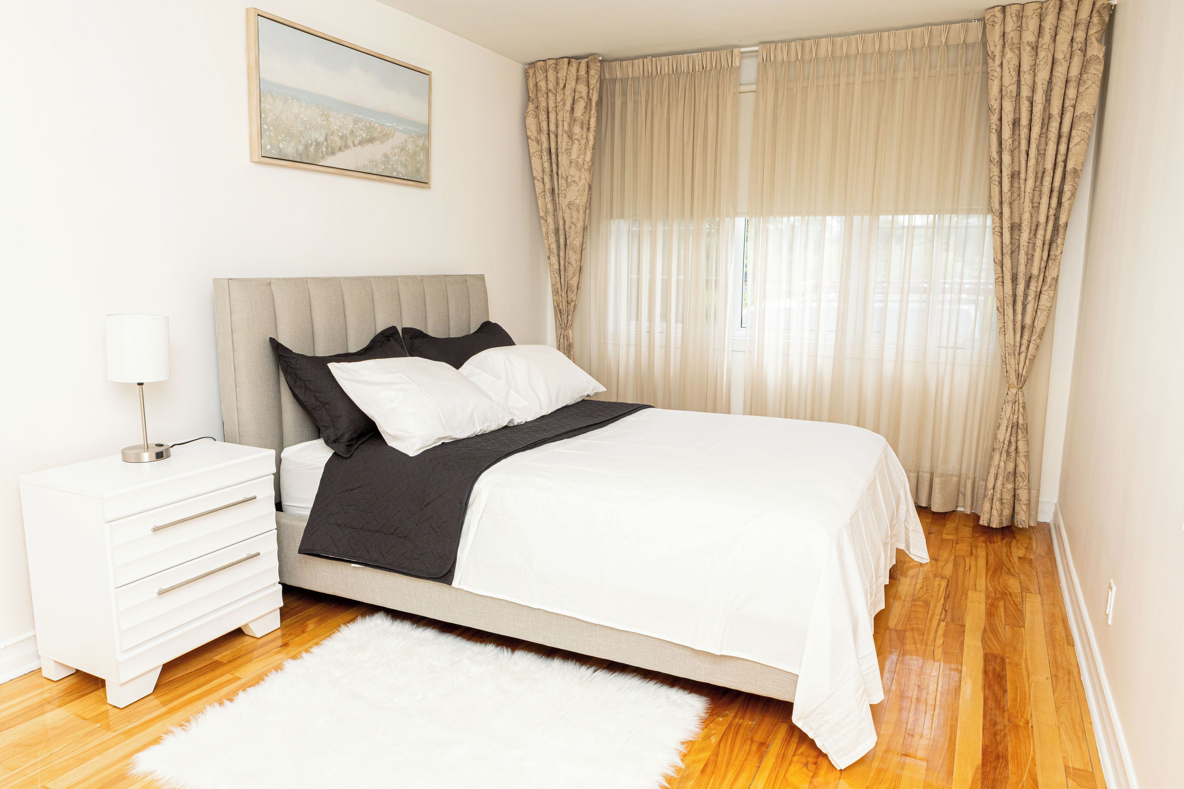 A bedroom with a bed, a nightstand, a lamp, a white rug, and a window with curtains.