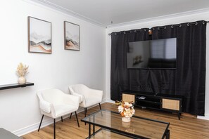Ein modernes Wohnzimmer mit einem Flachbildfernseher, zwei weißen Sesseln, einem Glastisch und Kunstwerken an der Wand.