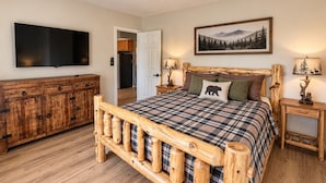 Quarto com estrutura de cama de madeira, televisão fixada na parede, cómoda de madeira e um quadro sobre a cama.
