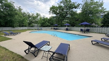 Piscina con sillas de descanso y sombrillas, rodeada de una valla y árboles.