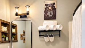 Uma casa de banho com toucador de madeira, sanita, um contentor de lixo, suporte de toalhas com toalhas, espelho e uma decoração de parede com tema de urso.