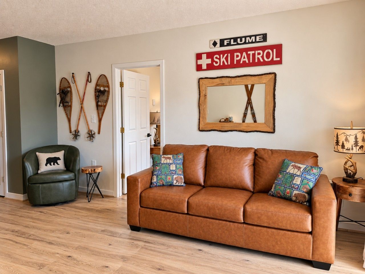 A living room with a brown leather sofa, two pillows with animal prints, a wooden coffee table, and a wall-mounted sign reading 'FLUME SKI PATROL'.