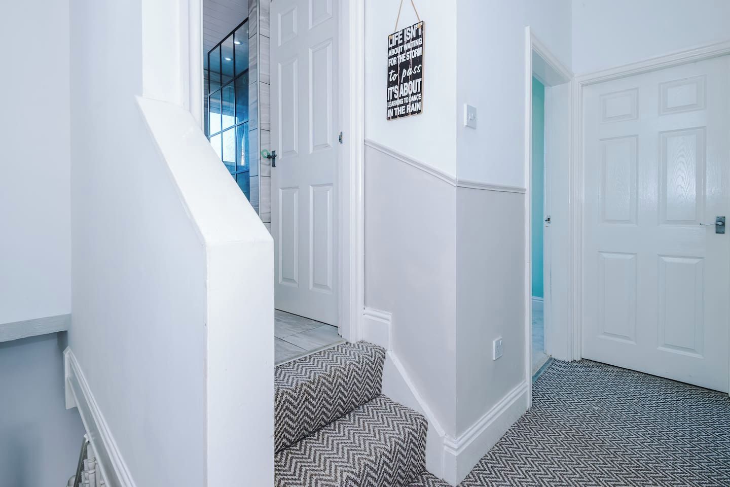 A hallway with white walls, a patterned carpet, a staircase with a herringbone design, and a door with a sign on the wall.