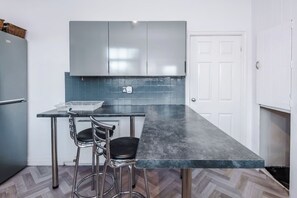 Cocina compacta con encimera oscura, dos taburetes tipo bar y una puerta blanca.