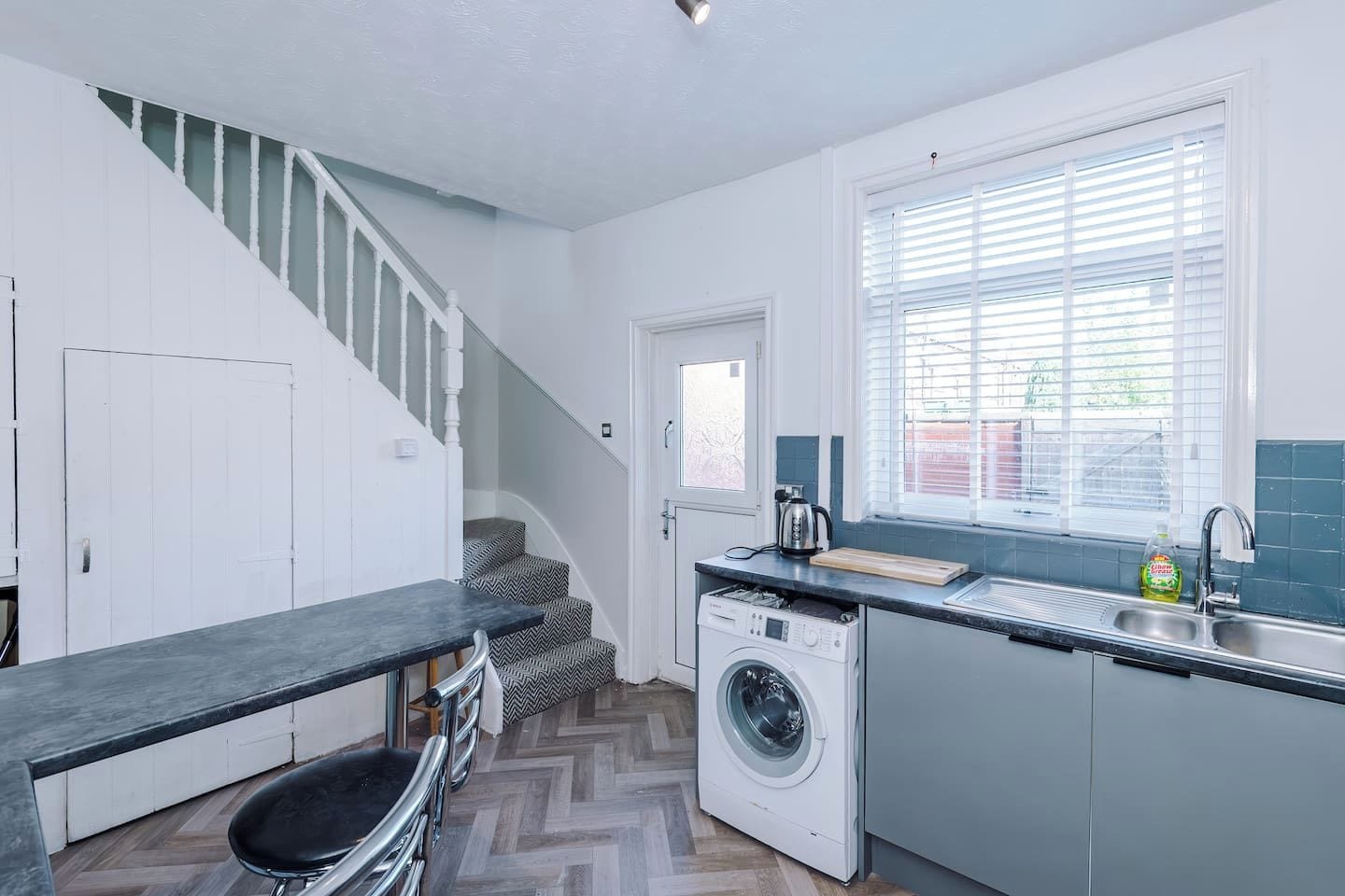 A modern kitchen with a built-in washing machine, a dining area with a bench, and a staircase leading to another level.