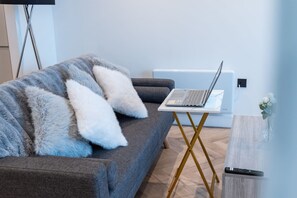 A modern living room with a grey sofa, a laptop on a white table, and a remote control on a wooden side table. - Central 1BR | Private Balcony | Weekday Discount (Greater Manchester)