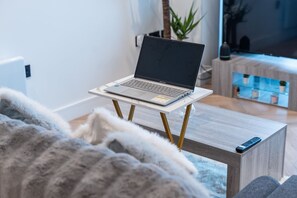 A laptop on a white table with wooden legs, a grey sofa, and a remote control on a wooden side table. - Central 1BR | Private Balcony | Weekday Discount (Greater Manchester)