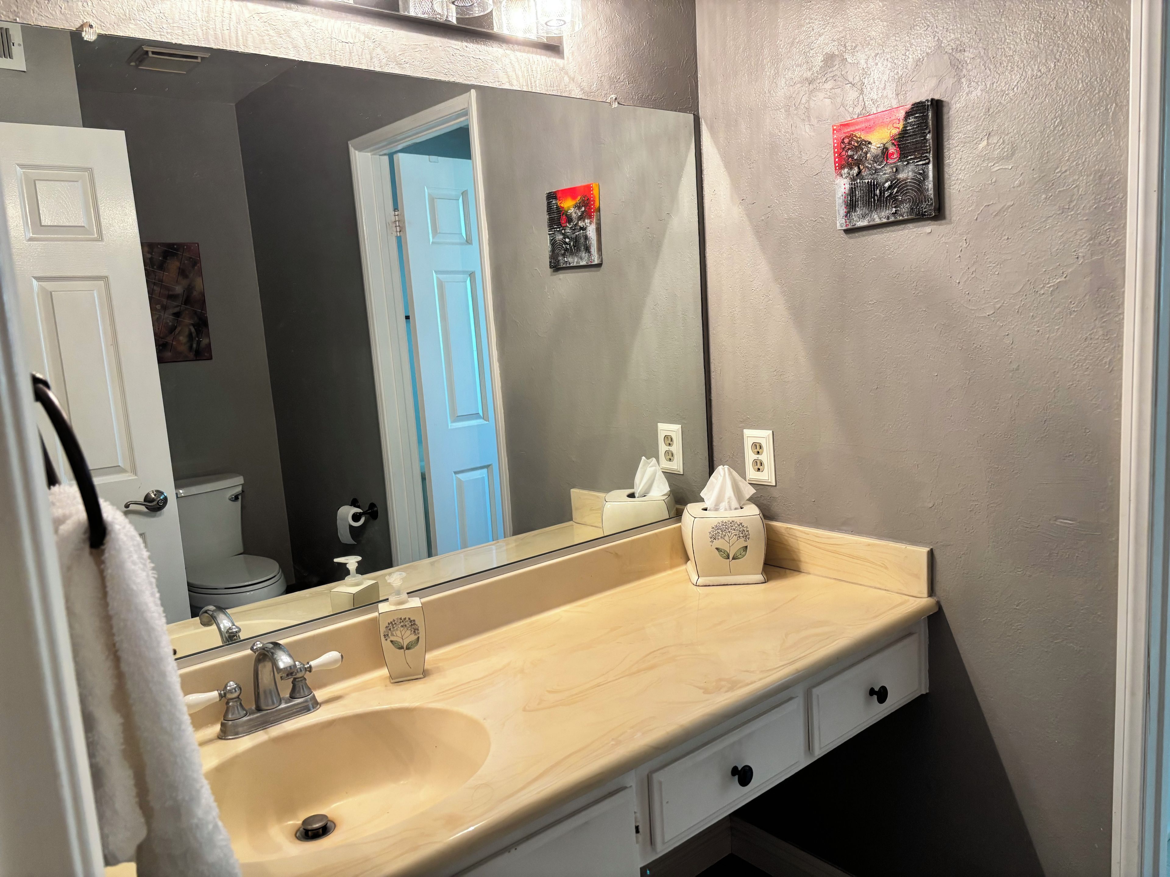 A bathroom with a beige sink, a mirror, a towel rack, and a toilet visible through an open door.
