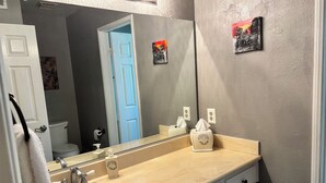 Ein Badezimmer mit einem beigefarbenem Waschbecken, einem Spiegel, einem Handtuchhalter und einer Toilette, die durch eine offene Tür sichtbar ist.