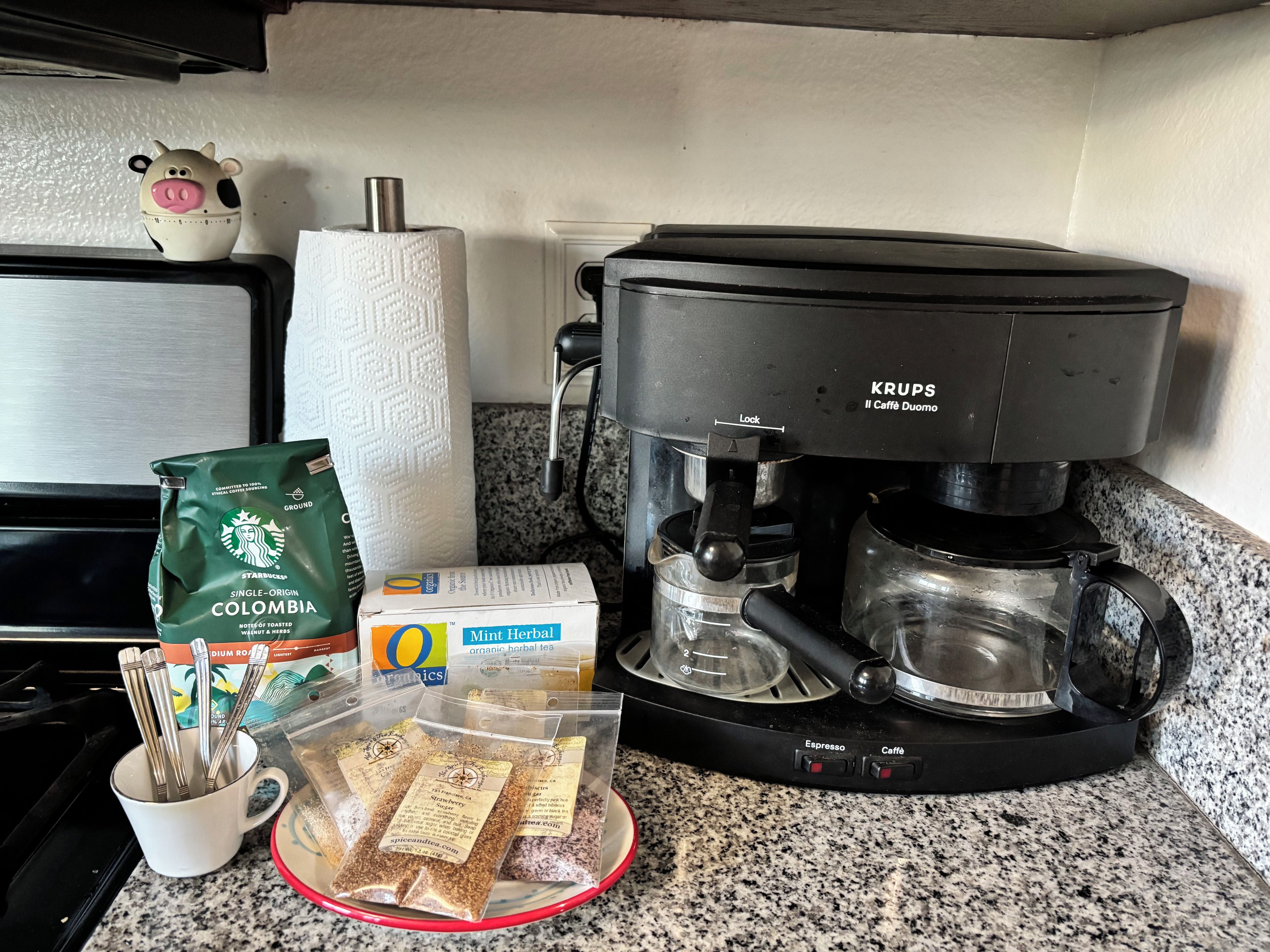 Eine Kaffeemaschine, eine Packung Starbucks Kaffee, eine Karton Oatly Hafermilch, ein Beutel Pfefferminztee und eine Tasse mit Besteck auf einer Küchenarbeitsfläche.