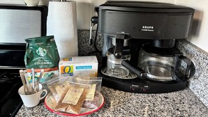 Eine Kaffeemaschine, eine Packung Starbucks Kaffee, eine Karton Oatly Hafermilch, ein Beutel Pfefferminztee und eine Tasse mit Besteck auf einer Küchenarbeitsfläche.