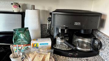 Eine Kaffeemaschine, eine Packung Starbucks Kaffee, eine Karton Oatly Hafermilch, ein Beutel Pfefferminztee und eine Tasse mit Besteck auf einer Küchenarbeitsfläche.