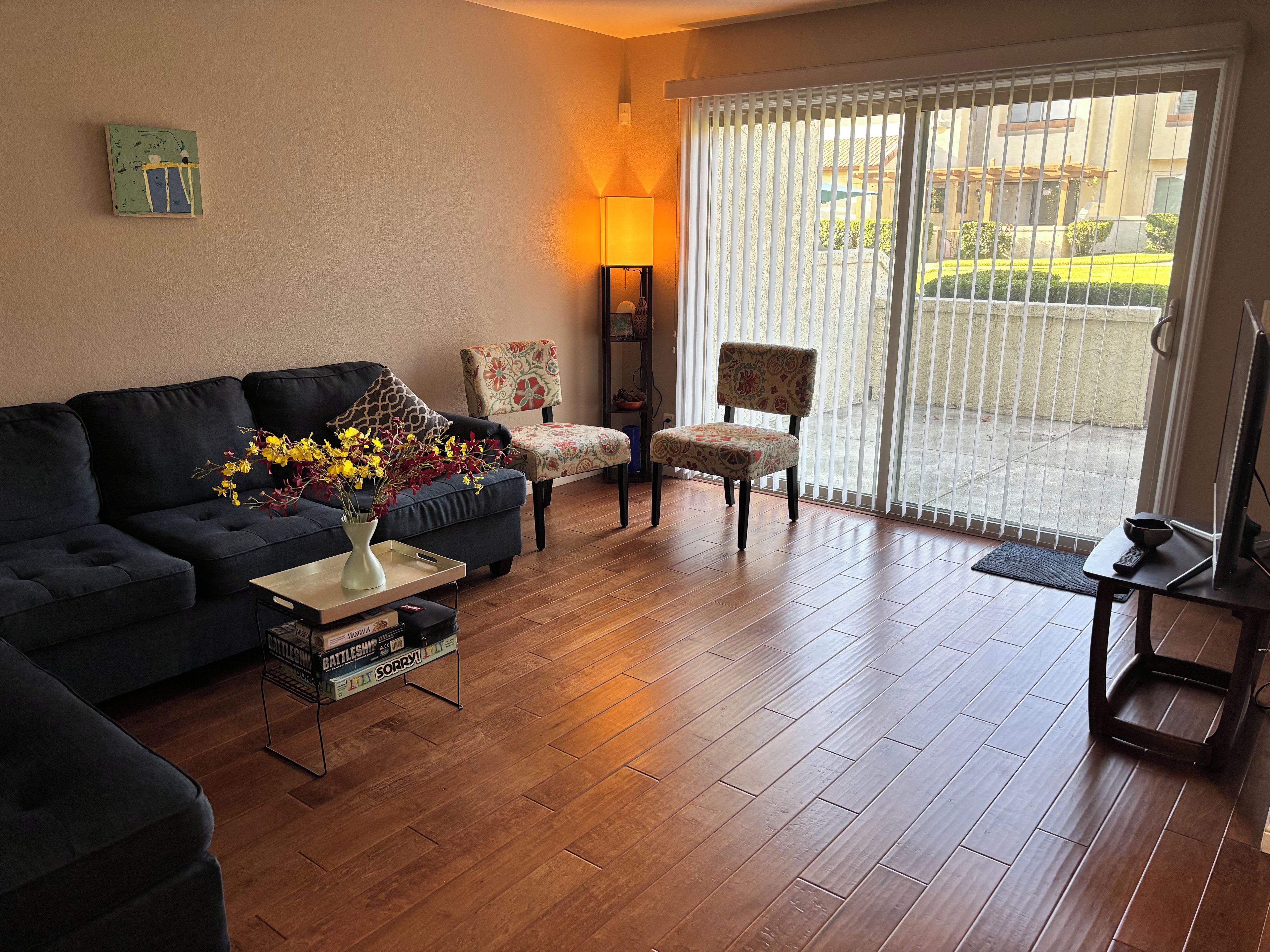 Ein Wohnzimmer mit einem dunkelgrauen Sofa, einem gemusterten Sessel, einem Couchtisch mit Blumenvase und einem Fernseher auf einem Schrank.