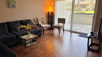 Ein Wohnzimmer mit einem dunkelgrauen Sofa, einem gemusterten Sessel, einem Couchtisch mit Blumenvase und einem Fernseher auf einem Schrank.