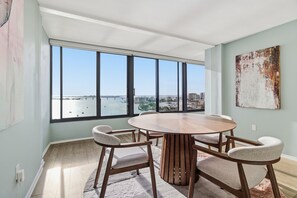 En matplats med ett runt bord och fyra stolar, stora fönster med utsikt över en stad och vatten, samt ett konstverk med en intressant yta på väggen.