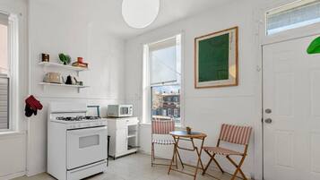 Une cuisine compacte équipée d’une cuisinière, d’un four à micro-ondes et d’étagères contenant des ustensiles de cuisine. On y trouve également un coin repas avec une table et des chaises, ainsi qu’une porte sur laquelle est posé un objet vert.