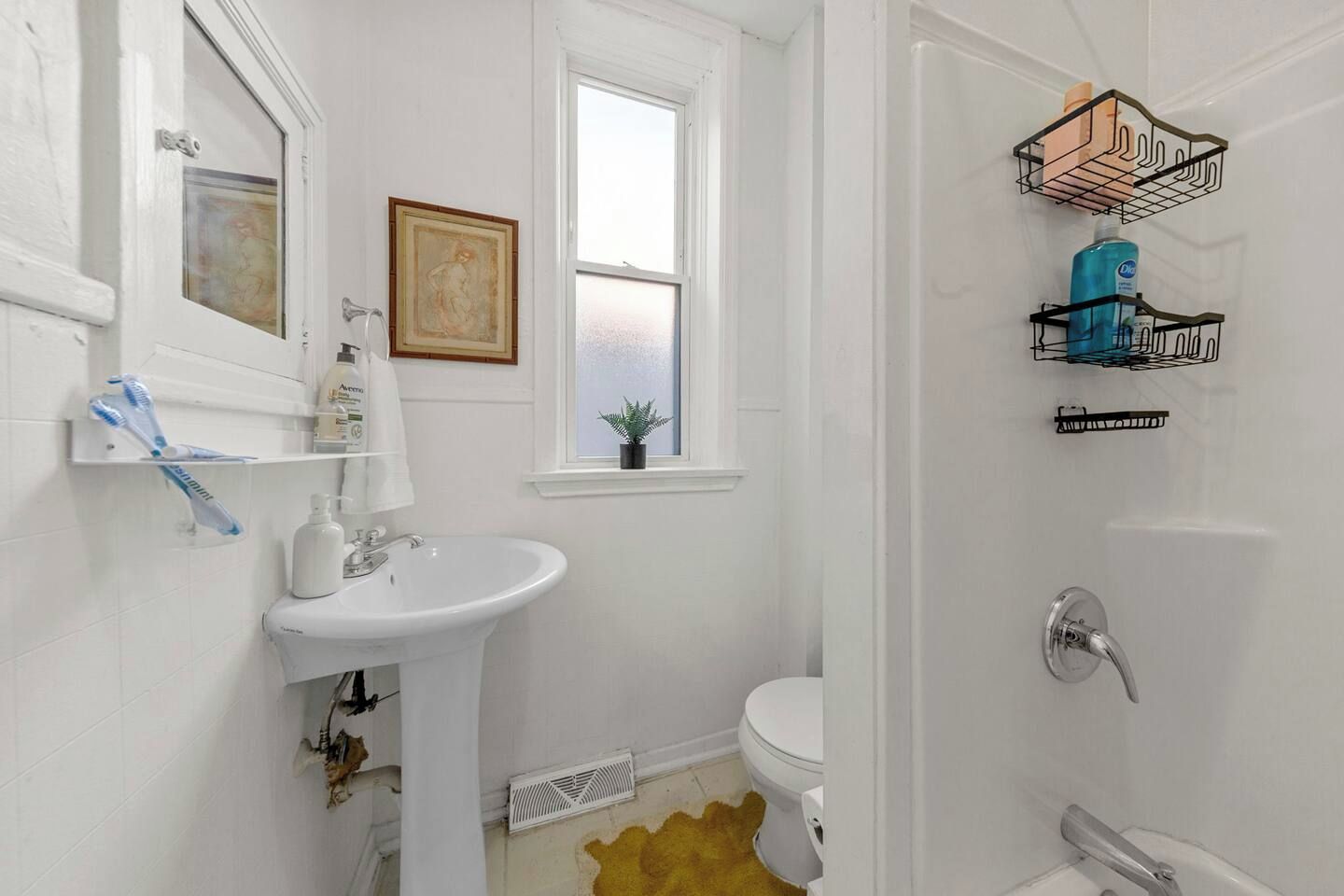 A bathroom with a white sink, toilet, and shower area. There are two wall-mounted shelves with toiletries and a framed picture above the sink.