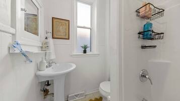 A bathroom with a white sink, toilet, and shower area. There are two wall-mounted shelves with toiletries and a framed picture above the sink.