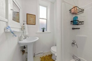 Ein Badezimmer mit weißem Waschbecken, WC und Duschbereich. Zwei Wandregale beherbergen Toilettenartikel, darüber hängt ein gerahmtes Bild.