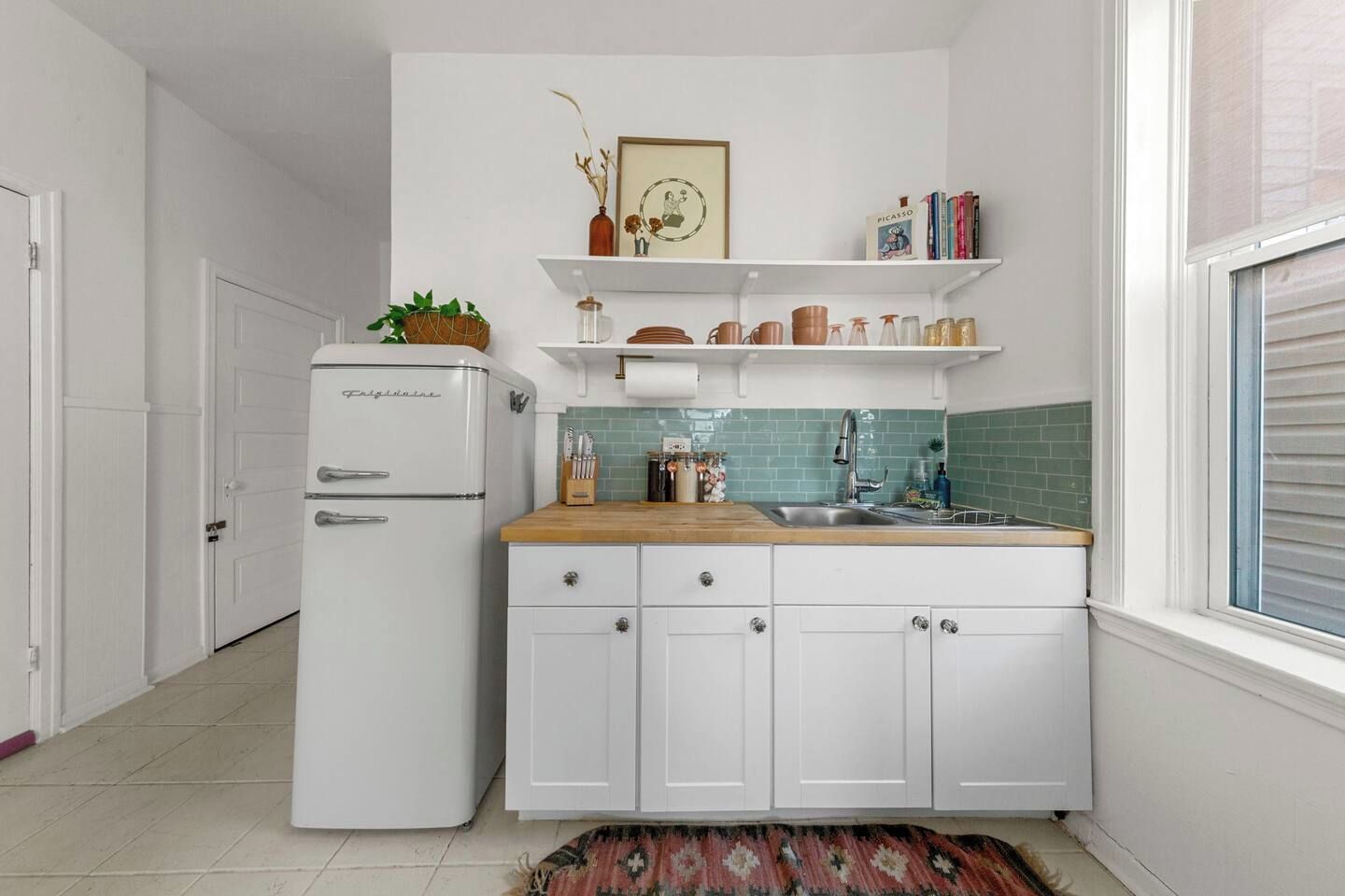 Een kleine keuken met witte kasten, een geïntegreerde koelkast, een houten aanrechtblad en een raam met zonwering.