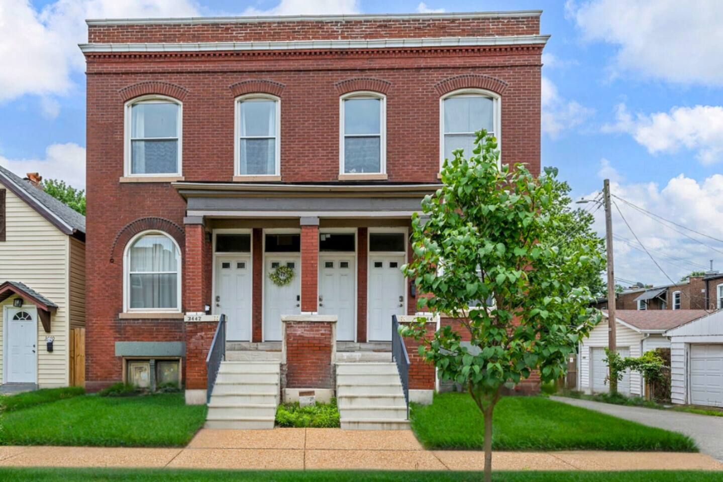 A two-story brick house with a front porch, a small tree in front, and a clear sky.