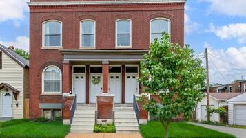 A two-story brick house with a front porch, a small tree in front, and a clear sky.