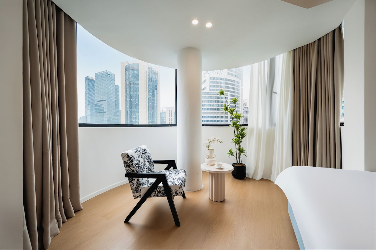 A modern hotel room with a curved ceiling, a large bed, a seating area with a chair, a small table with a vase and a plant, and a view of city buildings.