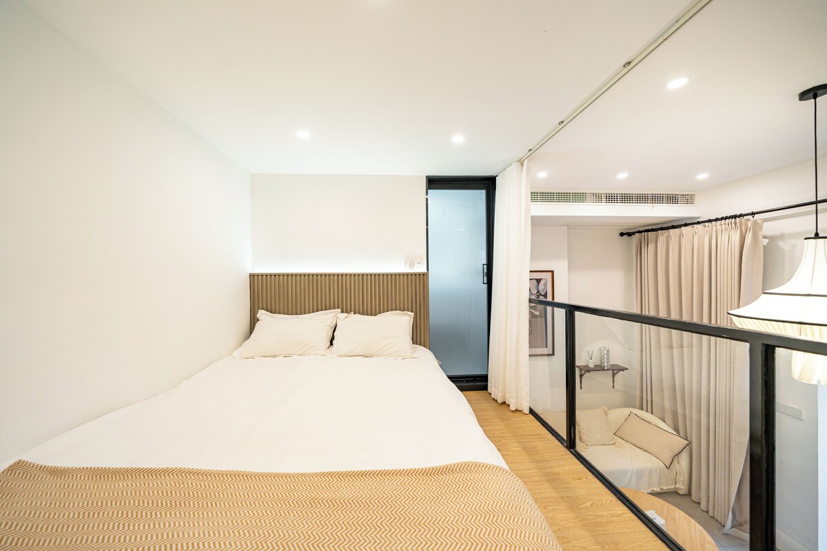 A modern bedroom with a large bed, a wooden headboard, a small seating area with a sofa, and a view of the outside.