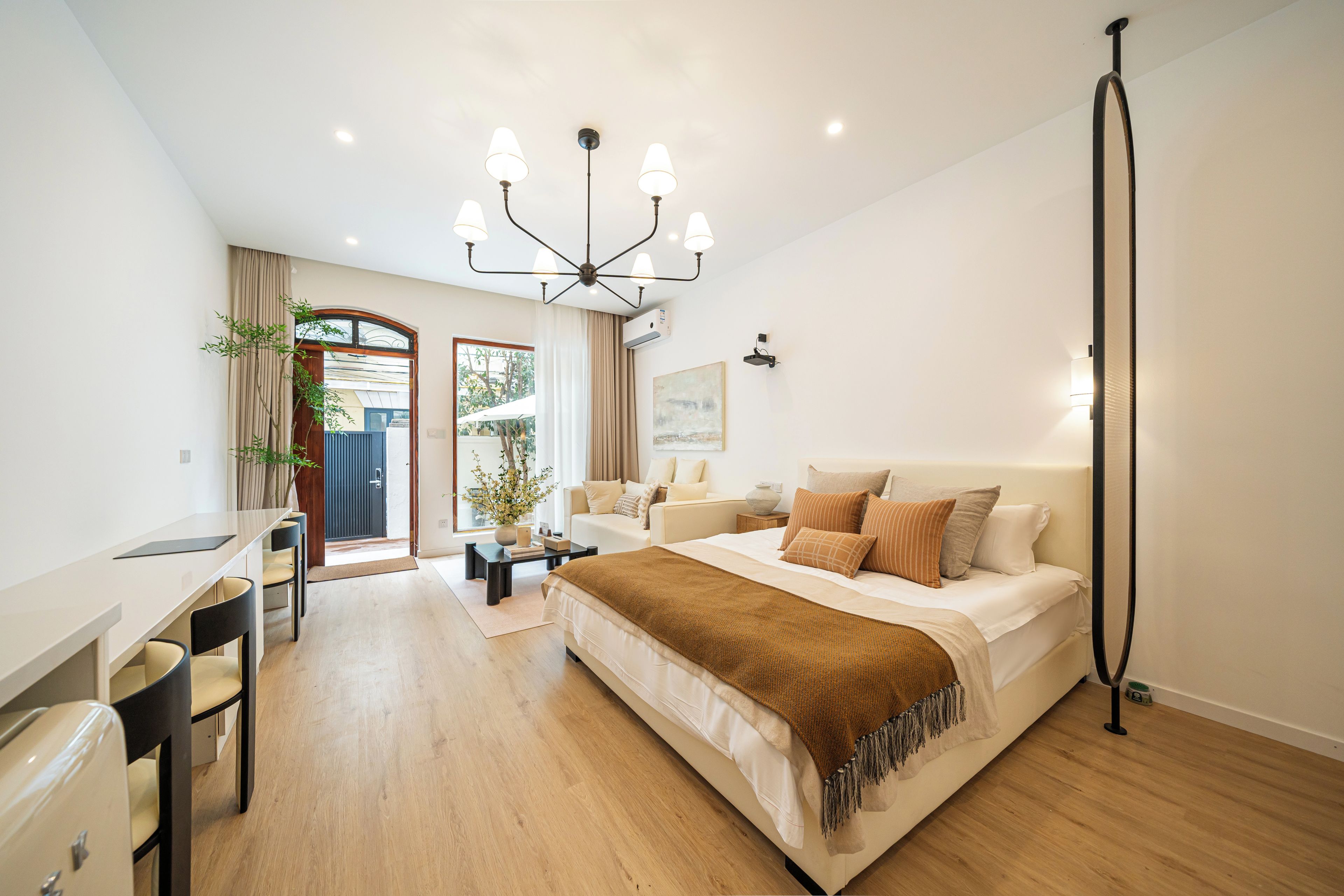 A modern bedroom with a large bed, a desk, and a chair. There is a window with curtains, a plant, and a ceiling light fixture.