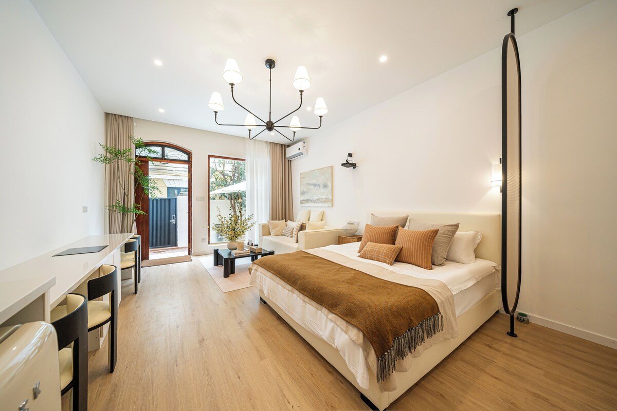 A modern bedroom with a large bed, a desk, and a chair. There is a window with curtains, a plant, and a ceiling light fixture.