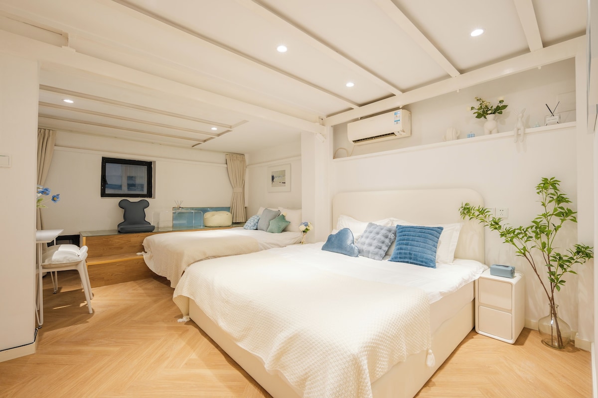 A hotel room with two beds, a desk, a chair, a small table, a plant, and an air conditioning unit.