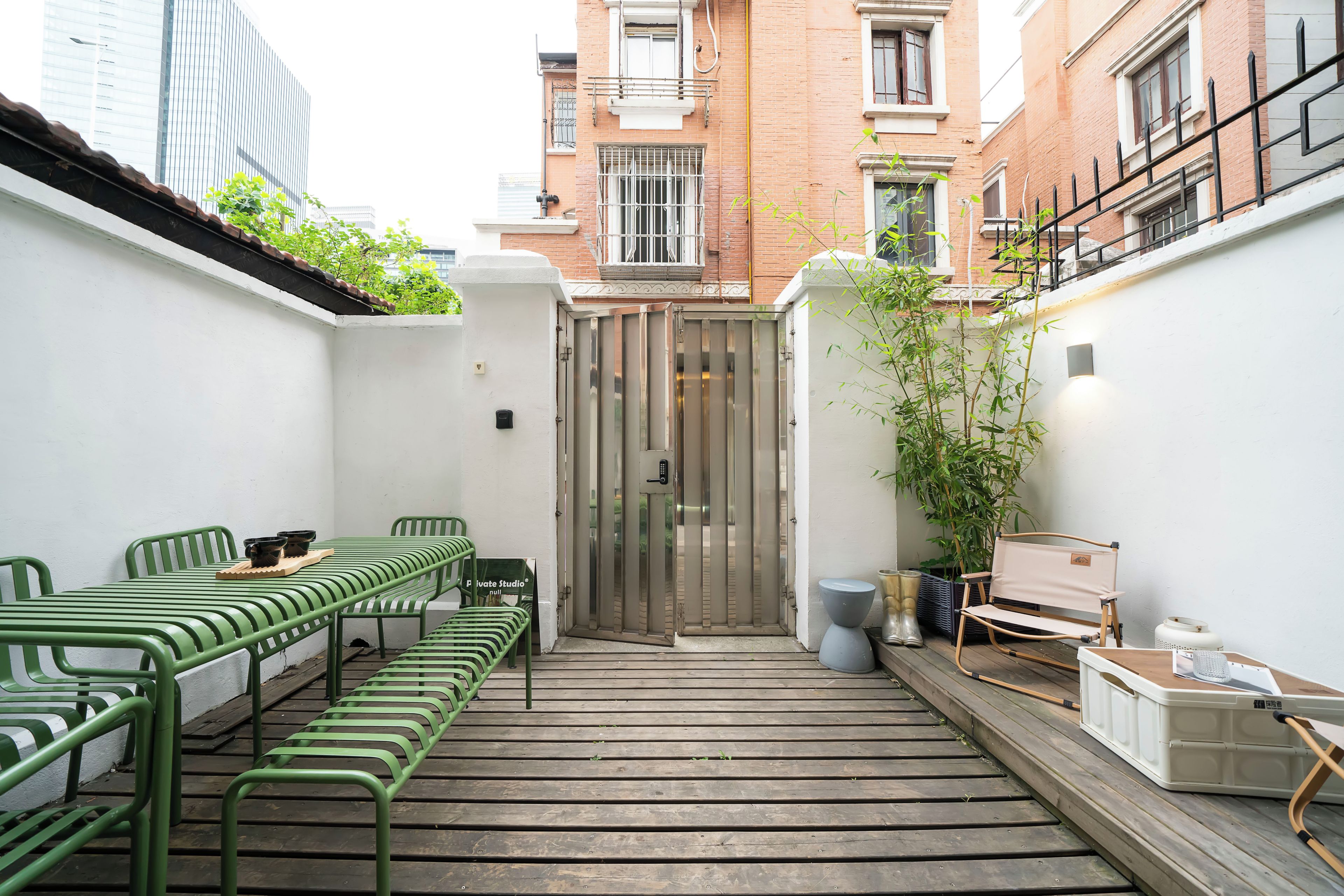 A narrow courtyard with a wooden deck, green metal chairs, a table with a tray, and a white wall with a door.