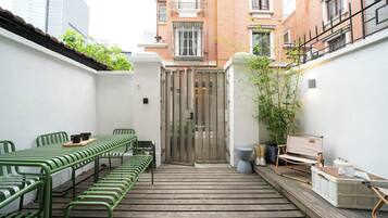 A narrow courtyard with a wooden deck, green metal chairs, a table with a tray, and a white wall with a door.