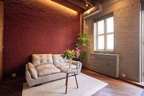 Une pièce avec un mur en briques, un radiateur, une fenêtre, un canapé et une plante posée sur une petite table.