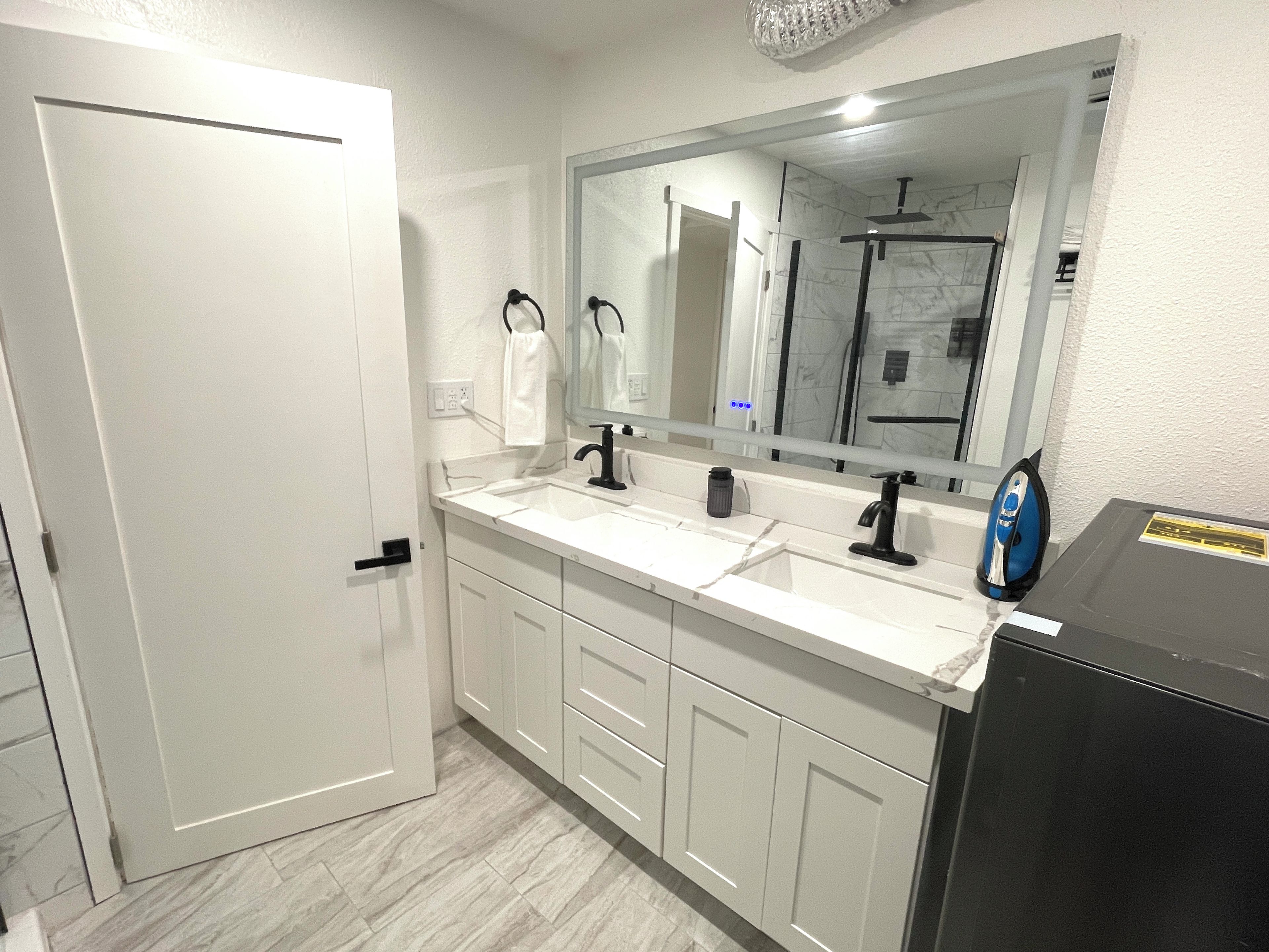 A bathroom with a white door, a double sink with black faucets, a large mirror, and a black cabinet.