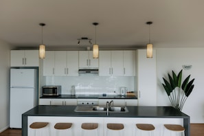 A modern kitchen with white cabinets, a stainless steel refrigerator, a microwave, a stove, and a sink. There are three pendant lights hanging above the island. - Oracle Beach House @ Penneshaw, Kangaroo Island (Penneshaw)