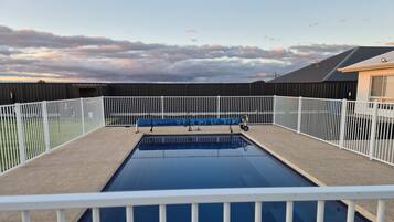 A rectangular swimming pool with a white fence surrounding it, set against a backdrop of a cloudy sky.