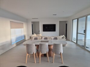 A modern dining room with a large table and chairs, a sofa, a flat-screen TV, and large windows.
