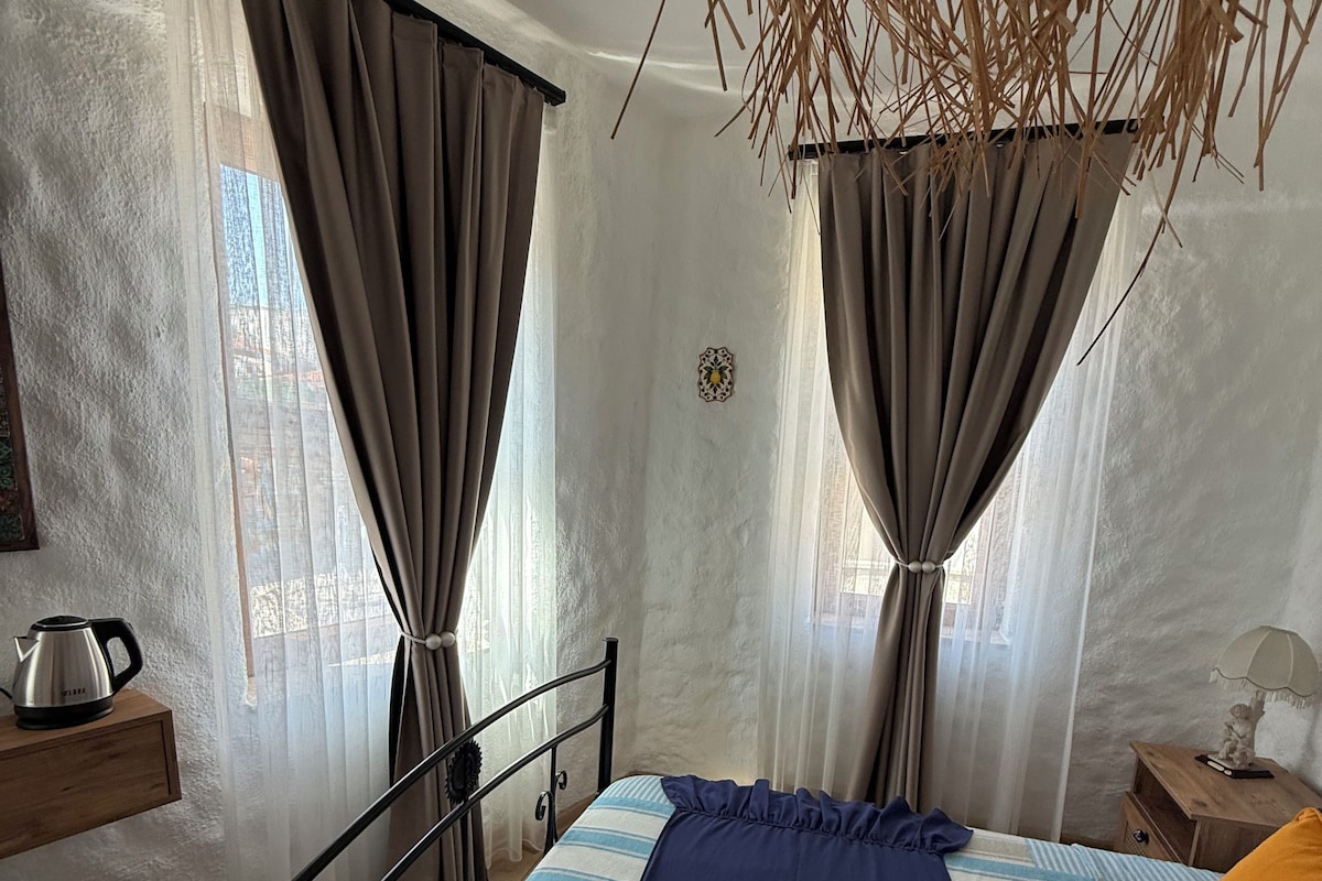 A bedroom with a bed, a nightstand, a wooden shelf, a window with curtains, and a hanging plant.