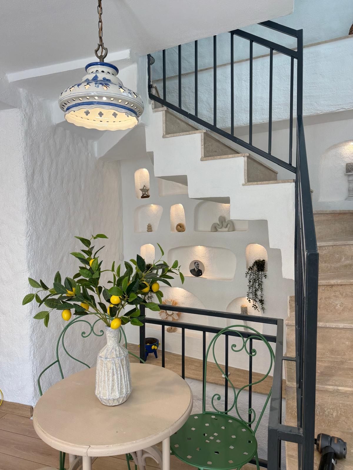 Un escalier avec une rampe noire, un mur blanc orné d’éléments décoratifs et une table sur laquelle est posé un vase rempli de citrons.