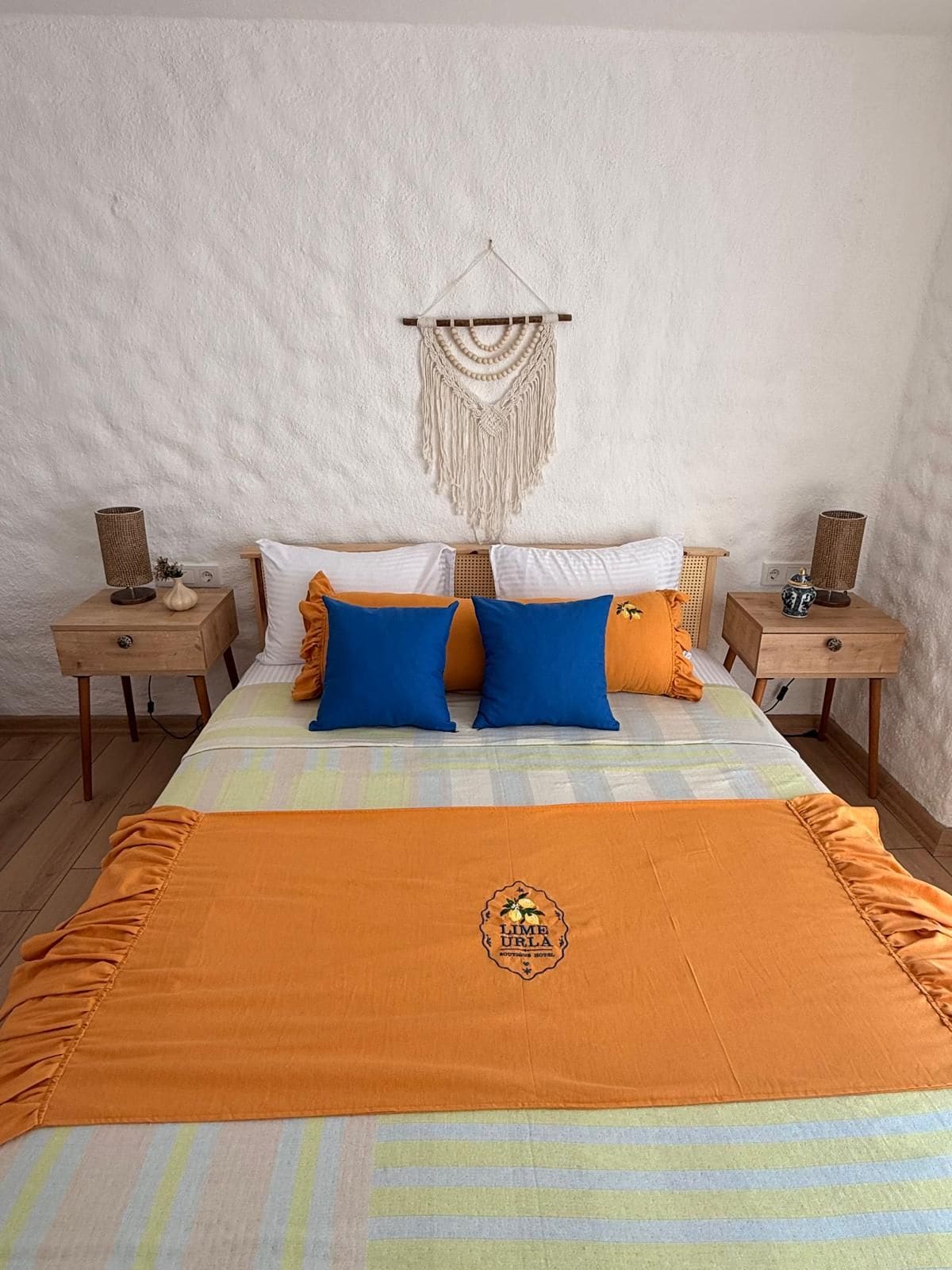 A bed with an orange blanket, two blue pillows, and a macrame wall hanging.
